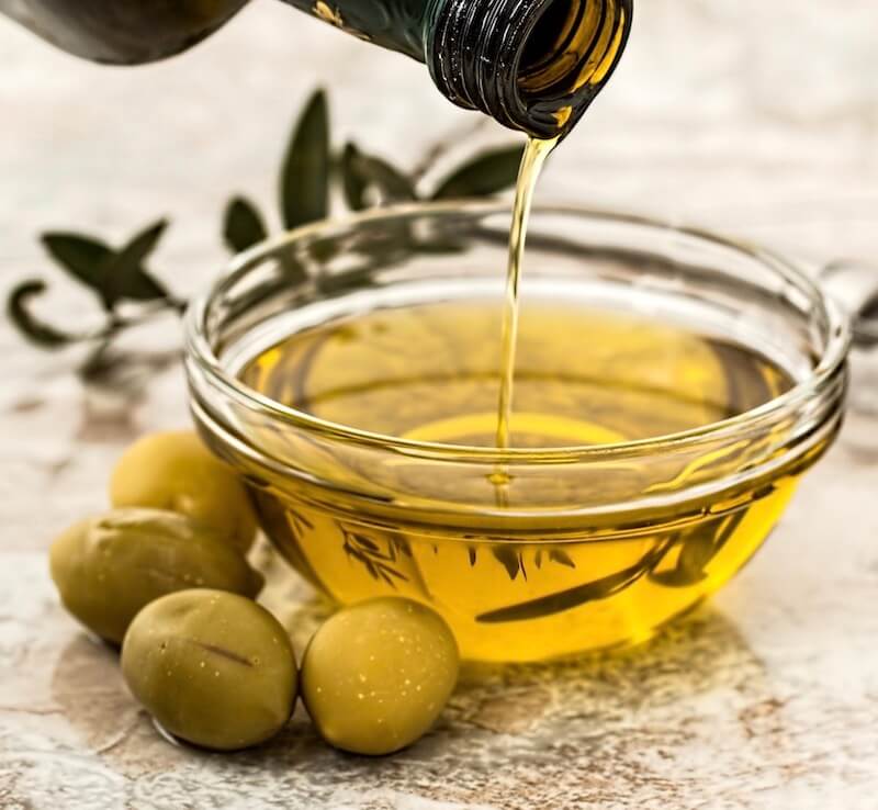 Olivenöl wird von einer Flasche in eine Schale gegossen, daneben liegen Oliven und ein Olivenzweig